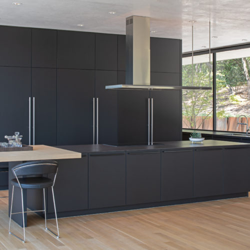 alt="View of full kitchen showing slate black matte satin lacquer cabinets, integrated table top in natural oak, and room-in-room walk-in pantry"