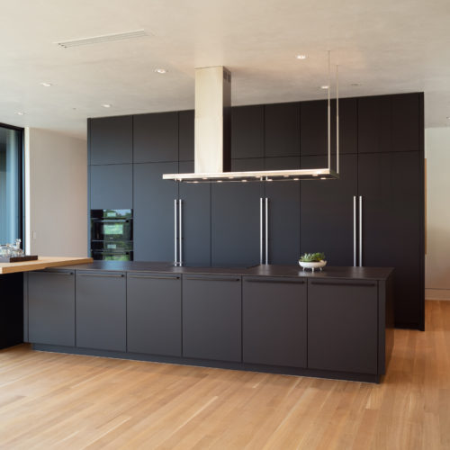 alt="Corner view of full kitchen showing slate black matte satin lacquer cabinets, gray metal handles, and natural oak integrated table top"