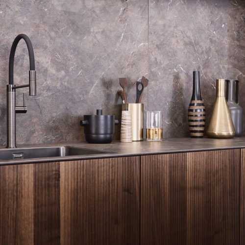 alt="side view of NASCA backsplash and worktop, steel faucet, and partial view of KERA cabinets with Walnut BOSSA fronts"
