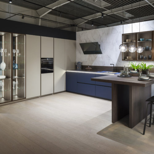 alt="corner view of full kitchen showing the VERO glass cabinets, a closed unit run that transition to the midnight blue handle-less run with inset lighting"