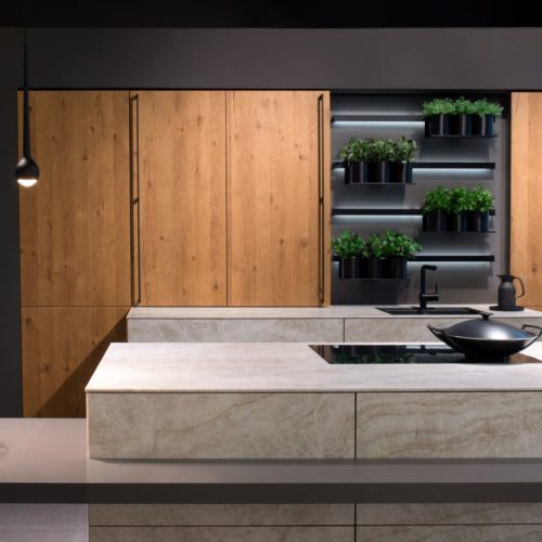 alt="Full frontal view of kitchen with double island with ROCCA fronts, matte dark gray table extension, wall cabinets with MADERO light oak veneers, and inset shelves with hanging plants and hidden LED strip lighting"