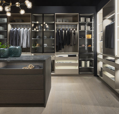 alt="Walk-in closet with matte lacquer cabinetry, VERO glass closets with strip LED lighting, and island with wood paneling"