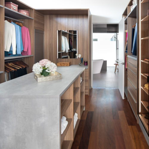 alt="Full view of primary walk in closet showing textured walnut laminate, inset LED strip lighting, concrete effect paneling, and gray integrated strip handles"